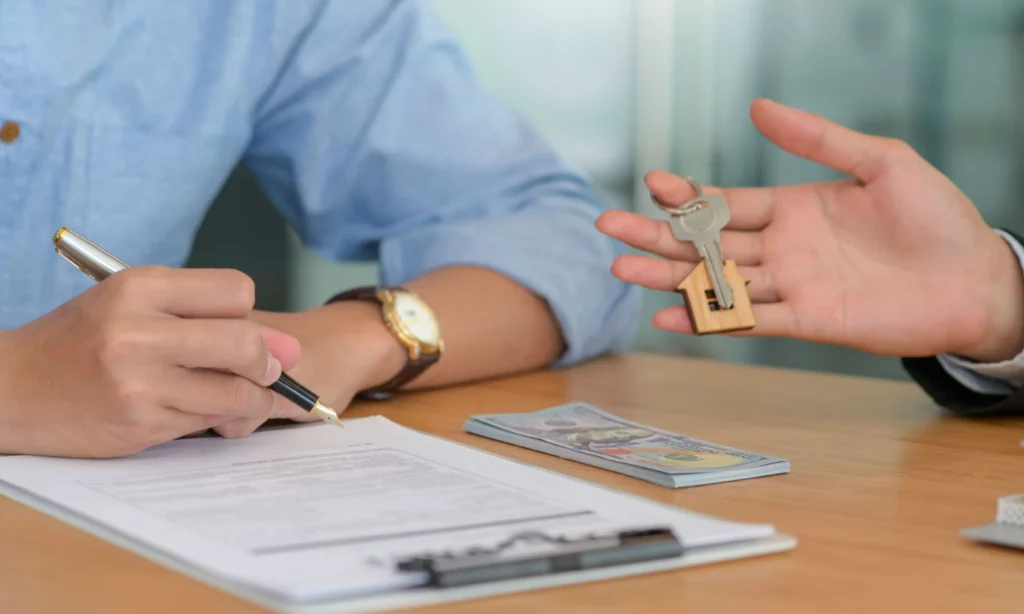 Person signs rental agreement while another hand holds house keys over a table with cash, representing a security deposit transaction