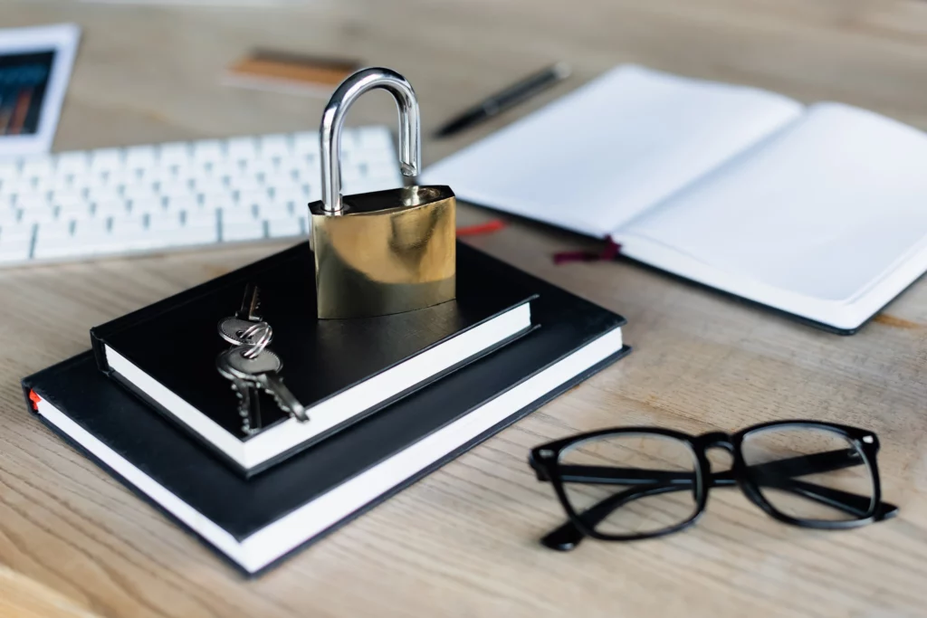 Locked padlock on stacked notebooks with keys, open notebook, eyeglasses, pen, and keyboard on a tidy desk.
