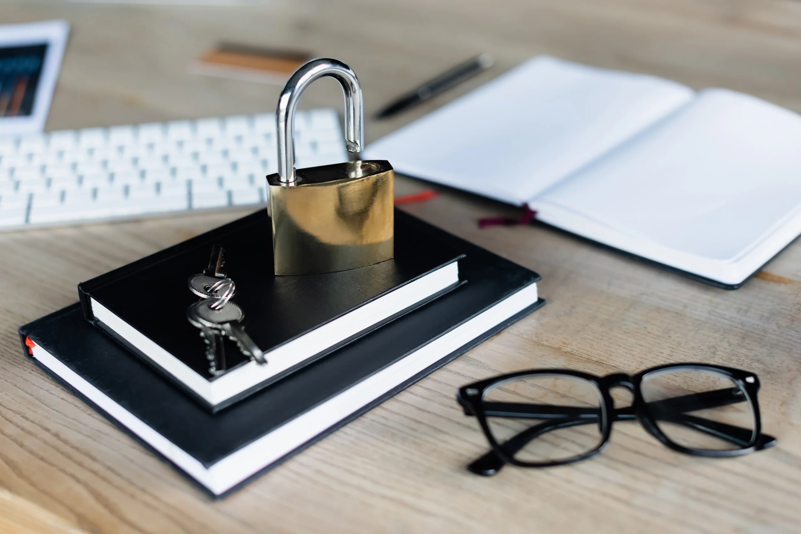 Locked padlock on stacked notebooks with keys, open notebook, eyeglasses, pen, and keyboard on a tidy desk.
