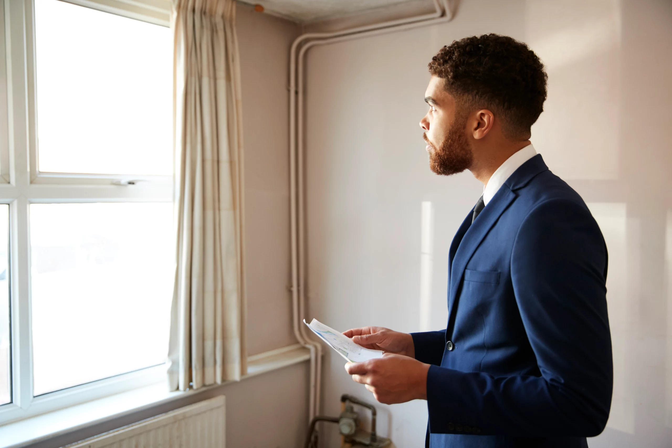 Man in blue suit holding papers, looking out window in thoughtful mood — considering rental property liability risks